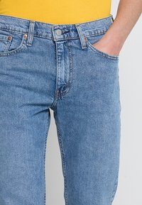 Personne portant un jean en denim bleu clair, avec une main dans la poche avant droite et un haut jaune visible à la taille.