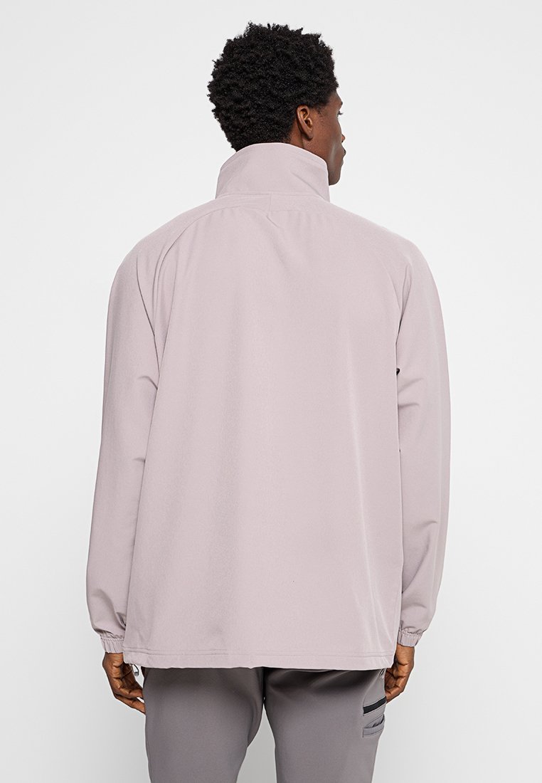 Mann in einer hellrosa Jacke mit hohem Kragen und grauen Hosen, der mit dem Rücken zu einer schlichten weißen Wand steht.