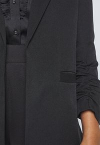 Černý šitý blazer s texturovanou látkou, jednou kapsou a nařasením na rukávu pro zvýšení designového zájmu.