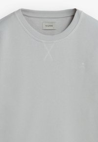 Sudadera gris claro con cuello redondo acanalado, que presenta un logotipo bordado en la esquina inferior derecha y un detalle de punto X en el cuello.