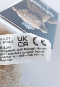 Clothing tag showing certification marks UKCA and CE, product details, and eco-friendly note, attached to beige fabric with blurred background.