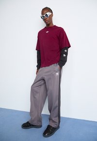 Bordeauxkleurig T-shirt met geborduurd logo, gelaagd over een zwart langemouwen T-shirt met sterpatroon. Grijze joggingsbroek met zijstrepen en zwarte schoenen.