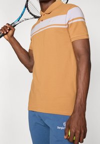 Brązowa koszulka polo w białe i pomarańczowe paski, z krótkim rękawem i logo na piersi. Model trzyma rakietę tenisową i piłkę.