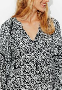 Blouse florale noir et blanc avec un col en V, un col froncé et des manches longues. Tissu texturé avec des garnitures noires et des détails à cordon.