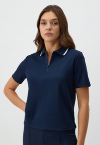 Donkerblauwe polo met korte mouwen en een halflange ritssluiting bij de kraag, met een witte streep langs de kraag. Glad textuur van de stof.