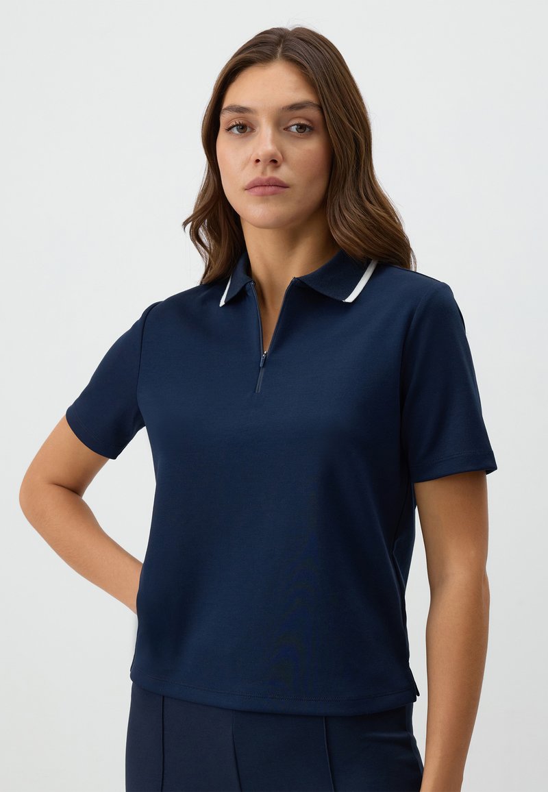Donkerblauwe polo met korte mouwen en een halflange ritssluiting bij de kraag, met een witte streep langs de kraag. Glad textuur van de stof.