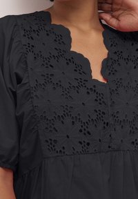 Musta mekko, jossa on kukkakuvioista pitsiä kauluksessa, aaltoilevat reunat ja teksturoitu kangas draping bodice kanssa.