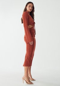 Ein rostfarbendes Kleid mit langen Glockenärmeln und seitlichen Ausschnitten. Verfügt über geraffte Details und eine taillierte Silhouette bis zur Mitte der Wade.