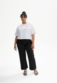 Witte cropped t-shirt met een klein rood grafisch ontwerp, gecombineerd met los zittende zwarte jeans met rechte pijpen en grijze sandalen. Eenvoudige, casual stijl.