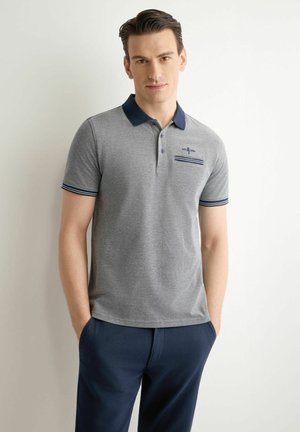 Mężczyzna w szarej koszulce polo z granatowym kołnierzem i mankietami, granatowe spodnie, stoi z rękami w kieszeniach na jednolitym białym tle.