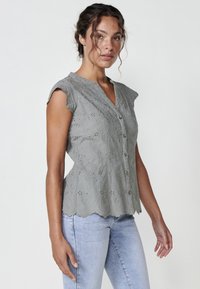 Hellgraue bestickte Bluse mit kurzen Ärmeln, gewelltem Saum, V-Ausschnitt und Blumendesign, knöpfbarer Front. Kombiniert mit hellen Jeans.