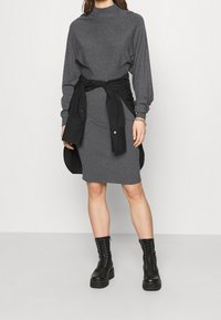 Grau gestreiftes Kleid mit langen Ärmeln, kombiniert mit einem schwarzen Shirt, das an der Taille gebunden ist. Schwarze Schnürstiefel verleihen einen robusten Akzent.