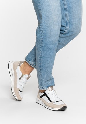 Osoba oblečená v světlých modrých džínách a bílých béžových teniskách s černými a kovovými akcenty, stojící na jedné noze na bílém pozadí.