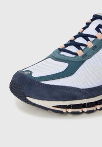 Sportos tornacipő eleje tengerészkék, türkiz, fehér hálós anyagból és bézs díszítésekkel, texturált talppal és keresztbe fűzött fűzőkkel, semleges háttér előtt.