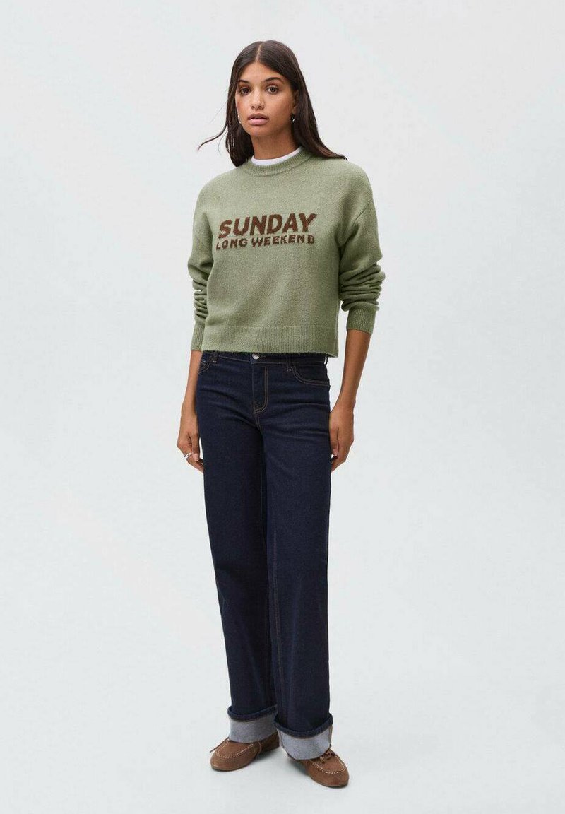 Zeleni pleteni pulover s natpisom "NEDJELJA DUGI WEEKEND", u kombinaciji s tamnoplavim širokim trapericama i smeškim mokasinama s podvrnutim rukavima.