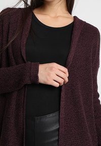 Cardigan en maille marron avec un tissu texturé, devant ouvert et manches longues. En dessous, un haut noir ajusté est partiellement visible.