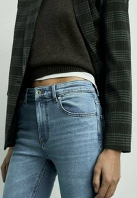 Jeans slim fit de culoare albastră, cu un efect de decolorare, având cinci buzunare, o textură din denim și un detaliu subtil de cusătură deschisă, asortați cu un pulover închis la culoare.