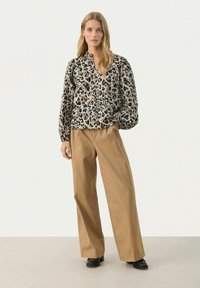 Bluse mit Leopardenmuster, hoher Kragen und Ballonärmeln, kombiniert mit lockeren beigefarbenen Hosen und schwarzen Schuhen vor einem neutralen Hintergrund.