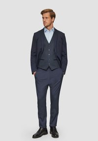 Marineblauw driedelig kostuum bestaande uit een getailleerd jasje, een gestreepte gilet en bijpassende broek, gecombineerd met een lichtblauw shirt en zwarte schoenen.