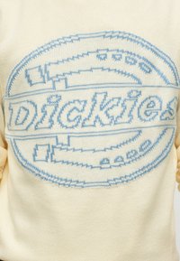 Pull crème avec grand logo Dickies bleu tricoté sur la poitrine dans un ovale avec des motifs stylisés de lame de scie.