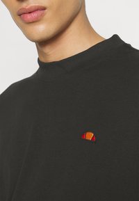 T-shirt noir en coton à manches courtes avec un col montant et un petit logo coloré brodé sur la poitrine. Texture lisse.