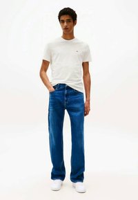Mladý muž stojící s rukama v kapsách, oblečený v bílém tričku, modrých džínách a bílých teniskách na bílém pozadí.