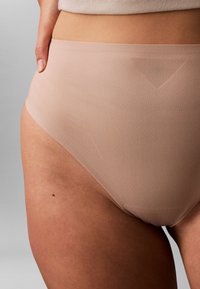 Nærbillede af en person iført glat, lysebeige sømløst undertøj med en ribbet talje og en lille modermærke på det øvre lår.