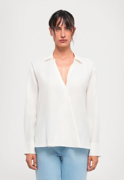 Kvinde med mørkt hår iført en hvid langærmet V-hals bluse og lyseblå jeans, stående mod en ensfarvet baggrund.