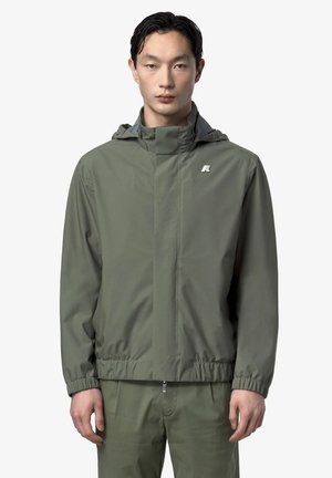 Giovane uomo che indossa una giacca con cappuccio verde oliva con cerniera e pantaloni abbinati, in piedi davanti a uno sfondo bianco uniforme.