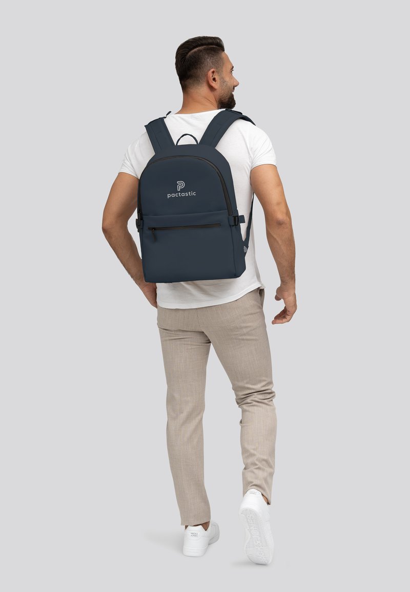 Zaino blu scuro con parte superiore arrotondata, tasca frontale con zip, spallacci regolabili e logo 'pactastic'. Texture liscia, design minimalista.