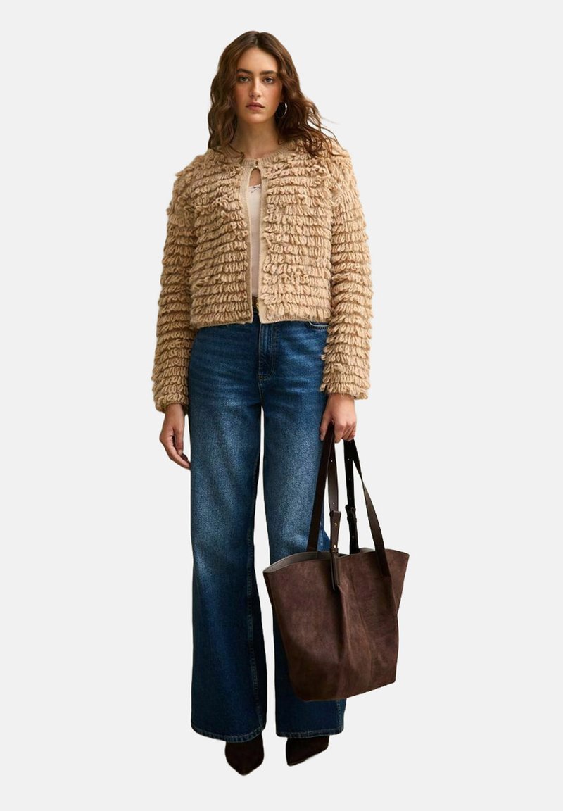Giacca beige texturizzata con finiture arricciate, indossata sopra un top chiaro, abbinata a jeans a zampa d'elefante blu e una borsa a spalla marrone scuro.