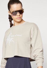 Avskuren beige sweatshirt med en grafisk soldesign i vitt. Modellen bär bruna solglasögon med tjocka, kantiga bågar.