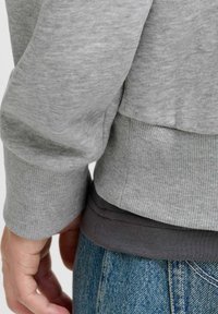 Šedá mikina s žebrovaným lemem, vrstvená přes tmavý top. Viditelné džíny z denimu se světlým praním a švy.