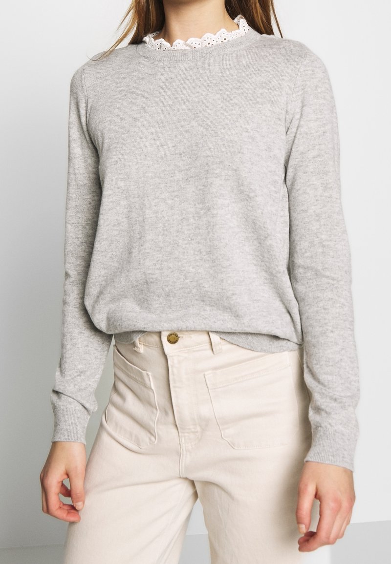 Femme portant un pull gris clair à manches longues avec un col en dentelle blanche et un pantalon taille haute écru, debout devant un fond uni.