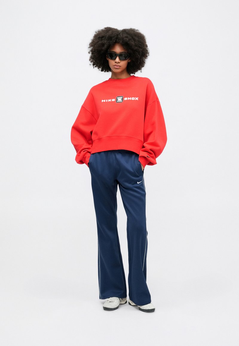 Felpa rossa over-size con logo "NIKE SHOX", abbinata a pantaloni blu con righe laterali. Sneakers bianche completano l'outfit.