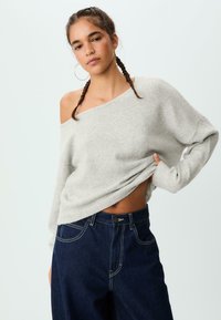 Hellgrauer, schulterfreier Strickpullover mit langen Ärmeln, der über eine strukturierte Oberfläche und einen gerippten Saum verfügt, kombiniert mit dunklen, hochtaillierten Jeans aus Denim.