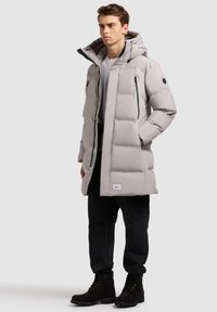 Svijetlo siva puffer jakna s kapuljačom, s crnim naglascima, zippered džepovima i oznakom blizu rubova. Nošena preko bijele majice i tamnih hlača.