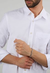 Bracciale d'argento con un design a catena elegante e una chiusura geometrica, indossato su un polso. La camicia è bianca, abbottonata e presenta una tasca.