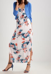 Floral maxi dress in white with blue, coral, and green patterns; side slits at hem. Paired with a light blue cardigan and high heel sandals.