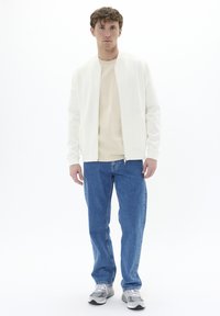 Chaqueta bomber blanca sobre una camiseta beige, combinada con jeans de denim azul y zapatillas grises. Diseño simple y limpio sin patrones visibles.