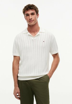 Joven con cabello rizado que lleva una camisa polo de punto blanca de manga corta y pantalones verde oliva, de pie relajado con una mano en el bolsillo.