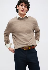 Beige kötött pulóver kerek nyakkal, világoszöld galléros ing fölött viselve, sötét nadrággal és barna övvel, amelynek arany csatja van.