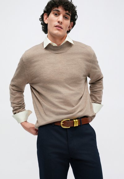 Beige adījuma sega ar apaļu apkakli, uzvilkta virs gaiši zaļas krekla ar apkakli, saskaņota ar tumšiem biksēm un brūnu jostu ar zelta sprādzi.