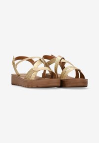 Sandales dorées avec des lanières croisées métalliques, semelle intérieure texturée marron et un talon compensé bas. Équipées de ferrures à boucle ajustables.