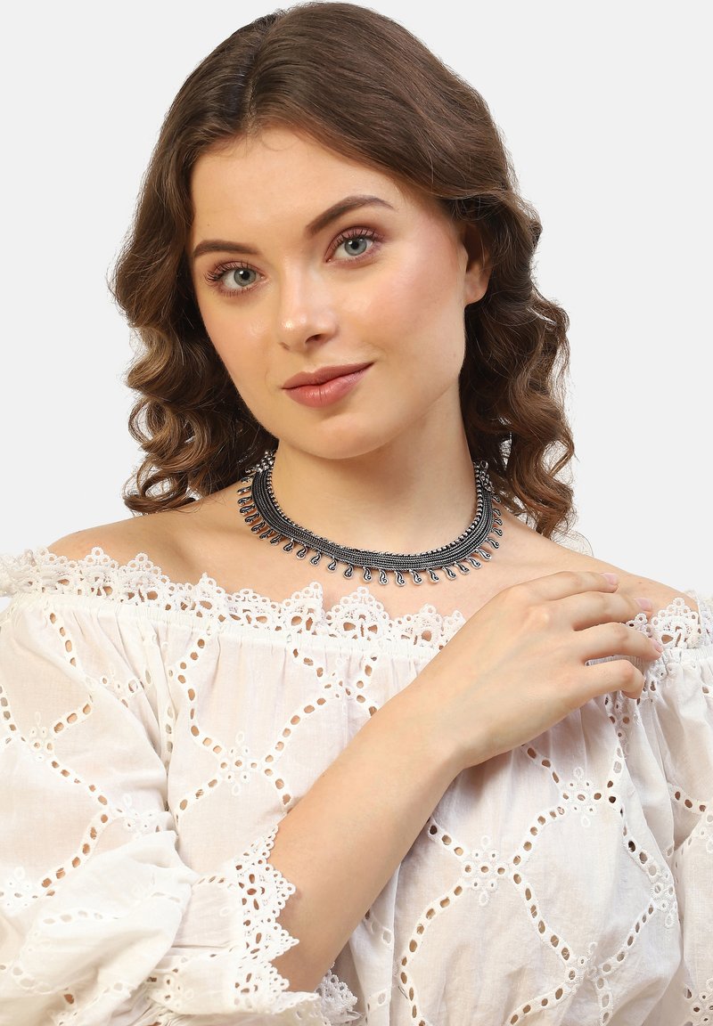 Chunky sølvfarvet halskæde med dekorative dråbeformede accenter, sat imod en model iført en hvid broderet off-shoulder top.