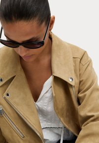 Hnědá semišová motocyklová bunda se stříbrnými zipovými detaily, nošená přes bílou strukturovanou blůzu, doplněná černými slunečními brýlemi.