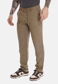 Pantaloni leggero marrone chiaro, comodi con vita regolabile con cordoncino, due tasche laterali e un tessuto a trama. Indossati con scarpe da ginnastica scure.