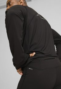 Juodas ilgomis rankovėmis sportinis marškinėlis su atspindinčiomis detalėmis, derinamas su juodomis leggings, turinčiomis užtrauktuką ir "DRYCELL" logotipą šalia liemens.