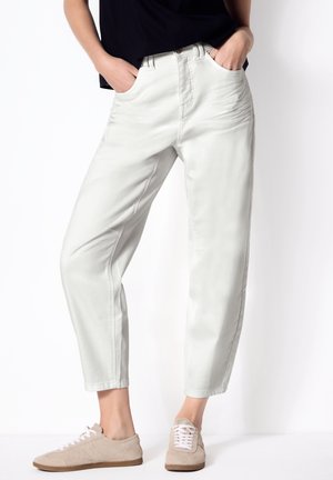 Persona che indossa pantaloni grigio chiaro a sigaretta e scarpe da ginnastica beige, in piedi con le mani nelle tasche davanti a uno sfondo bianco.