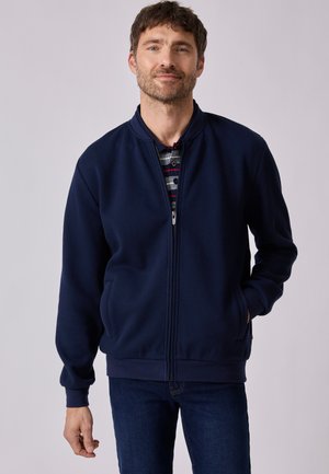 Uomo con capelli castani corti indossa una giacca blu navy con zip sopra una camicia a righe, in piedi con una mano in tasca davanti a uno sfondo semplice.